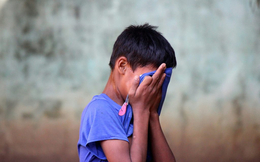 Boy dressed in blue t shirt with hands to his face as if crying into a towel