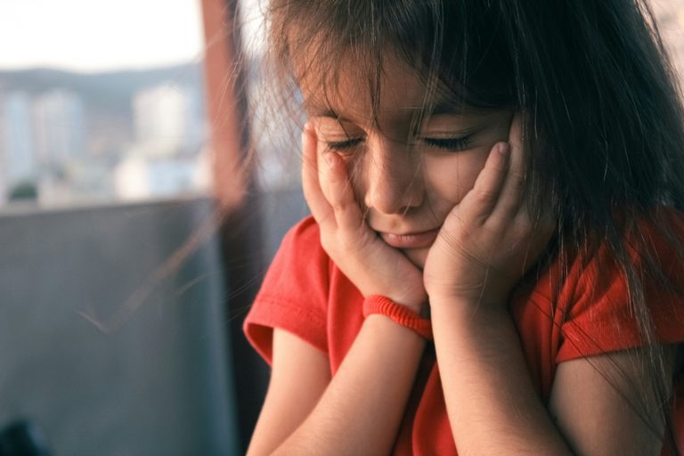 Girl in red t shirt holding face with both hands and eyes closed loking upset